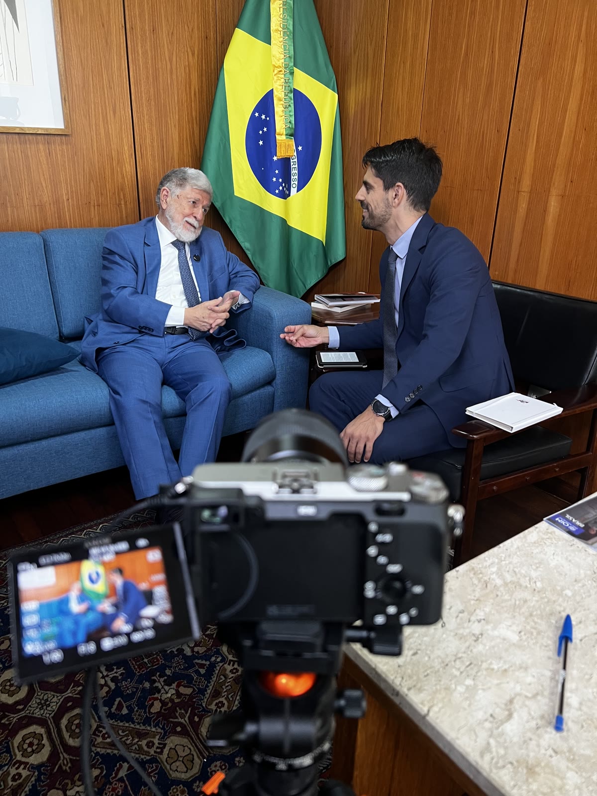 Entrevista institucional com bandeira do Brasil e câmera em primeiro plano